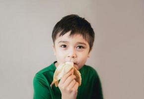 menino saudável comendo banana, criança feliz tomando café da manhã, criança bonita olhando para a câmera com um sorriso no rosto. foto