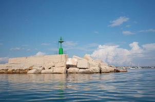 uma torre de sinalização de metal verde fica na costa do mar mediterrâneo. a torre fica em muitos blocos de pedra. ao fundo o céu azul refletido na água e nas ondas. foto
