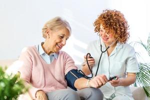 mulher sênior feliz tendo sua pressão arterial medida em um lar de idosos por seu cuidador. enfermeira feliz medindo a pressão arterial de uma mulher sênior na sala de estar foto