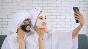 duas meninas asiáticas felizes em roupões de banho brancos com máscara facial tirando foto no smartphone juntos na sala de estar.