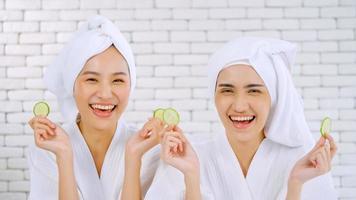 duas meninas asiáticas felizes em roupões de banho brancos com toalhas na cabeça segurando fatias de pepino na sala de estar. foto