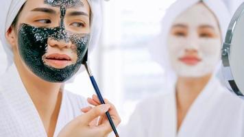 menina asiática em roupão branco, aplicando uma máscara preta revitalizante no rosto. foto