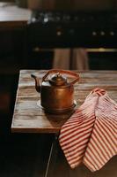 chaleira de alumínio retrô na mesa de madeira com toalha listrada vermelha perto. bule de cobre velho usa para fazer chá. utensílios de cozinha à moda antiga