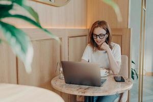 jovem e linda mulher ruiva freelancer em fones de ouvido trabalha remotamente no laptop no café foto