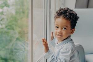 lindo menino de cabelos cacheados de raça mista com camisa azul clara ao lado da grande janela foto