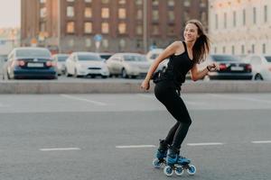 tiro ao ar livre de mulher alegre magro vestida com roupa ativa desvia o olhar leva passeios de estilo de vida saudável em poses de rolos na estrada em lugar urbano se sente energizado. patins como hobby. lazer foto