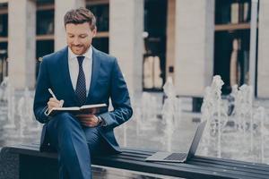 pensativo sorridente jovem empresário planejando o dia de trabalho, fazendo anotações no notebook ao ar livre foto