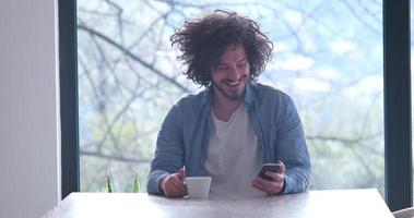 jovem tomando café e usando um celular em casa foto