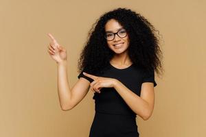 tiro isolado de mulher afro-americana feliz aponta para cima e para o lado, demonstra espaço de cópia em branco para o seu anúncio, usa óculos, camiseta casual, tem um sorriso gentil no rosto. pessoas e promoção foto