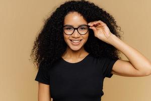 linda jovem afro-americana sorri para a câmera, mantém a mão na borda dos óculos, feliz em ouvir boas notícias, vestida com camiseta preta casual, isolada sobre fundo marrom. conceito de felicidade foto