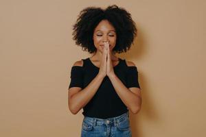 jovem afro-americana orando e de mãos dadas em gesto de oração em pé no estúdio foto