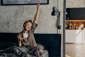 menina adorável relaxada com sono em pijama de cetim tentando acordar da noite de sono com café foto