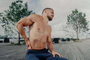 Desagradado desportista europeu com barba toca a parte inferior das costas e sente dor nas costas após exercícios de treinamento físico, poses ao ar livre. esporte exercitando lesão. cara atlético sofre dor, precisa de massagem foto