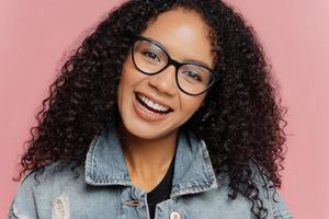 Feche o retrato de uma mulher sorridente feliz com penteado afro encaracolado escuro, inclina a cabeça, usa óculos ópticos e jaqueta jeans, isolada sobre fundo rosado. pessoas, etnia, emoções e sentimentos foto