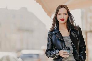 tiro ao ar livre do modelo feminino milenar atraente com maquiagem, vestido com jaqueta de couro elegante, bebe café do copo descartável, vagueia pela cidade, gosta de momentos de lazer. pessoas, bebendo conceito foto