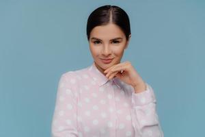 tiro na cabeça da linda mulher morena mantém a mão sob o queixo, tem cabelo penteado, maquiagem, pele saudável, usa camisa de bolinhas elegante, isolada sobre fundo azul. pessoas, beleza, conceito de feminilidade foto