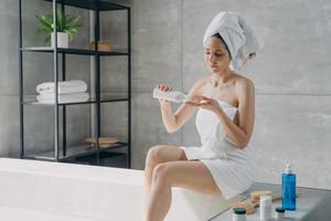 mulher europeia atraente envolta em toalha aplicando loção corporal após o banho. rotina diária de beleza.
