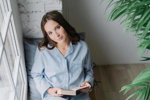 vista superior da mulher de aparência agradável relaxada gosta de ler livro, vestido com um terno doméstico casual, senta-se no peitoril da janela para boa luz e visão, gosta de solidão e tempo livre. conceito de hobby foto