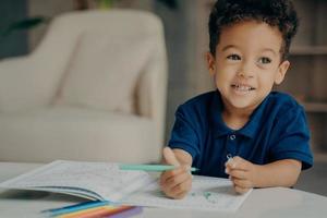 menino afro-americano bonitinho em camiseta polo azul escuro pintando livro de colorir em casa foto