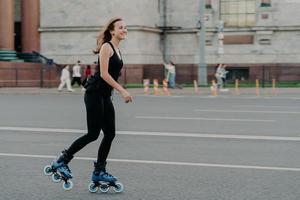 mulher atlética de patins se move ativamente posa no asfalto na rua vestida com roupas ativas tem expressão alegre. jovem modelo fitness patinando em local urbano. patins estilo de vida esportivo foto