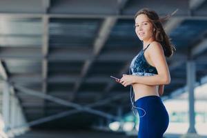 tiro horizontal de mulher atlética esbelta com figura perfeita, vestida com top casual e leggings, segura telefone celular moderno, ouve músicas populares em fones de ouvido, posa ao ar livre. pessoas e conceito de descanso foto