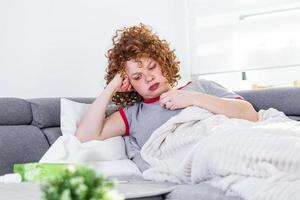 mulher fraca, vestindo roupas casuais está sentada em um sofá e tomando uma pílula. dia doente em casa. jovem tem resfriado comum. doença, conceito de problema de vírus sazonal. foto