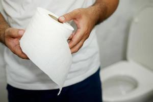 homem sofre de diarréia mão segure o rolo de papel de seda na frente do vaso sanitário. constipação no banheiro. tratamento dor de estômago e higiene, cuidados de saúde foto