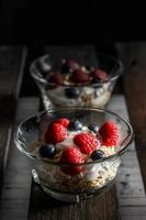 framboesas, mirtilos, cereais e iogurte em uma tigela de vidro em tábuas de madeira velhas. pequeno-almoço saudável para uma vida saudável. imagem vertical.