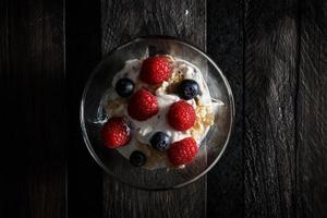 framboesas, mirtilos, cereais e iogurte em uma tigela de vidro em tábuas de madeira velhas. pequeno-almoço saudável para uma vida saudável. vista de imagem horizontal de cima. foto