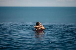 mulher feliz em maiô nadando na piscina infinita contra a beira-mar. foto