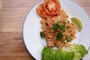 arroz frito de carne de porco e salsicha com cebola servido em um prato branco sobre uma mesa de madeira clara. foto