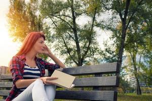 mulher ruiva pensativa relaxando no banco do parque com um livro. foto