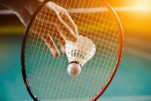 raquete de badminton e peteca branca velha segurando nas mãos do jogador enquanto servia pela rede à frente, desfocar o fundo da quadra de badminton e foco seletivo foto