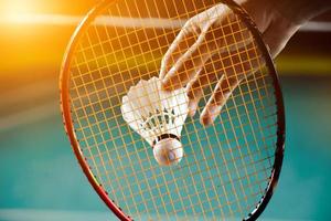 raquete de badminton e peteca branca velha segurando nas mãos do jogador enquanto servia pela rede à frente, desfocar o fundo da quadra de badminton e foco seletivo foto