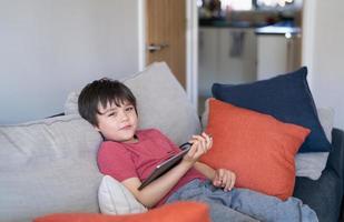 criança autêntica sentada no sofá assistindo desenhos animados ou jogando no tablet, menino criança usando a lição de aprendizagem de almofada digital na internet, educação em casa, conceito de educação on-line de ensino a distância foto