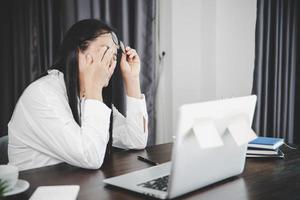 jovem mulher de negócios com sono e tem problemas oculares de frustração com a doença de cefalalgia por usar o computador portátil em sua mesa de escritório. funcionária estressada cansada e exausta por excesso de trabalho. foto