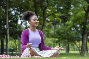 mulher afro-americana praticando relaxantemente meditação na floresta para alcançar a felicidade da sabedoria da paz interior para o conceito de mente e alma saudável