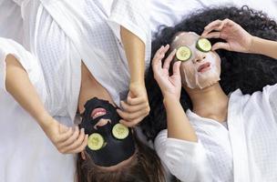 casal de namoradas em roupão fazendo rotina de cuidados com a pele usando máscara facial e fatia de pepino em férias de spa para pele de beleza e conceito de tratamento foto