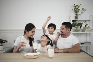 uma família tailandesa asiática saudável, crianças pequenas e pais jovens bebem leite branco fresco em vidro e pão alegria juntos em uma mesa de jantar de manhã, bem-estar nutrição em casa café da manhã refeição estilo de vida. foto