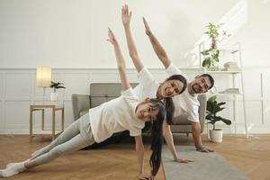 pais tailandeses asiáticos e filha exercício de treinamento de fitness e prática de ioga no chão da sala de estar, adorável remo juntos para saúde e bem-estar e estilo de vida doméstico feliz no fim de semana em família. foto