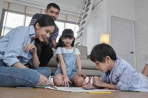 os irmãos e a mãe tailandeses asiáticos estão sentados no chão da sala, desenhando com lápis de cor juntos, o pai relaxando no sofá, atividade de fim de semana feliz adorável e estilo de vida doméstico de bem-estar. foto