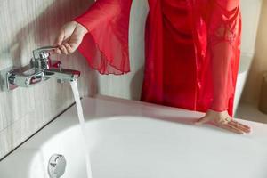 uma mulher de camisola de seda vermelha abre a torneira da banheira foto