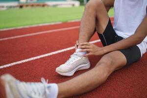 atletas esporte homem corredor vestindo branco sportswear sentado entorse de tornozelo sentindo dor no tornozelo ou perna ou joelho depois de praticar na pista de corrida no estádio, copie o espaço. conceito de lesão de esporte de corredor. foto