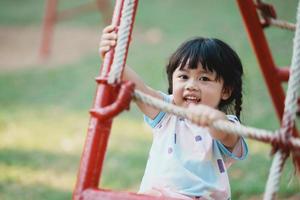 sorriso de linda garota asiática brinca na escola ou jardim de infância ou playground. atividade de verão saudável para crianças. menina asiática escalando ao ar livre no playground. criança brincando no playground ao ar livre. foto