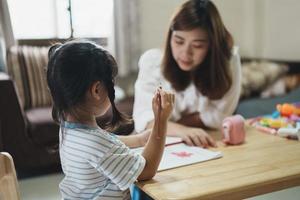 criança bonitinha pintando com tintas coloridas. menina asiática e mãe usando cor de desenho de giz de cera. filha e mãe fazendo lição de casa colorindo personagens de desenhos animados. conceito de estilo de vida de atividade de artista de bebê. foto