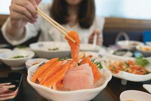 mulher asiática comendo um sashimi de salmão. mulher usando pauzinho para pegar sashimi de peixe cru da tigela branca. mulher usando pauzinhos fatiados de salmão cru, comida japonesa em um restaurante japonês. foto