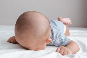 cabeça de bebê vestindo uma camisa listrada, rastejando em um colchão branco.