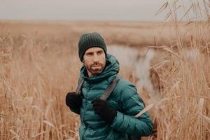 tiro de jovem bonito com barba por fazer escura, carrega mochila, olha para trás, percebe algo, tem expressão pensativa, passa o tempo livre ao ar livre. Tempo de outono. pessoas e conceito de aventura foto