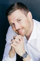 retrato vertical de homem bonito e alegre com barba por fazer com sorriso amigável, mantém as palmas das mãos pressionadas sob o queixo, usa relógio na mão, olha alegremente para a câmera. trabalhador jovem profissional foto