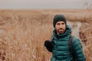 tiro horizontal de homem bonito contemplativo tem barba por fazer, usa chapéu, jaqueta e luvas, fica perto do fundo do campo de trigo com espaço livre no lado direito. conceito de pessoas, lazer e caminhada foto